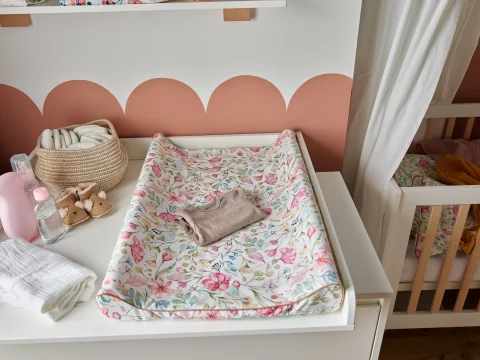 Lifestyle'owe zdjęcie białej komody z szufladami marki Pinio z kolekcji Floral, na blacie znajduje się przewijak pokryty tekstylną matą w kwiatowy wzór, mebel w kolorze białym, standardowy rozmiar, aranżacja w pokoju niemowlęcym