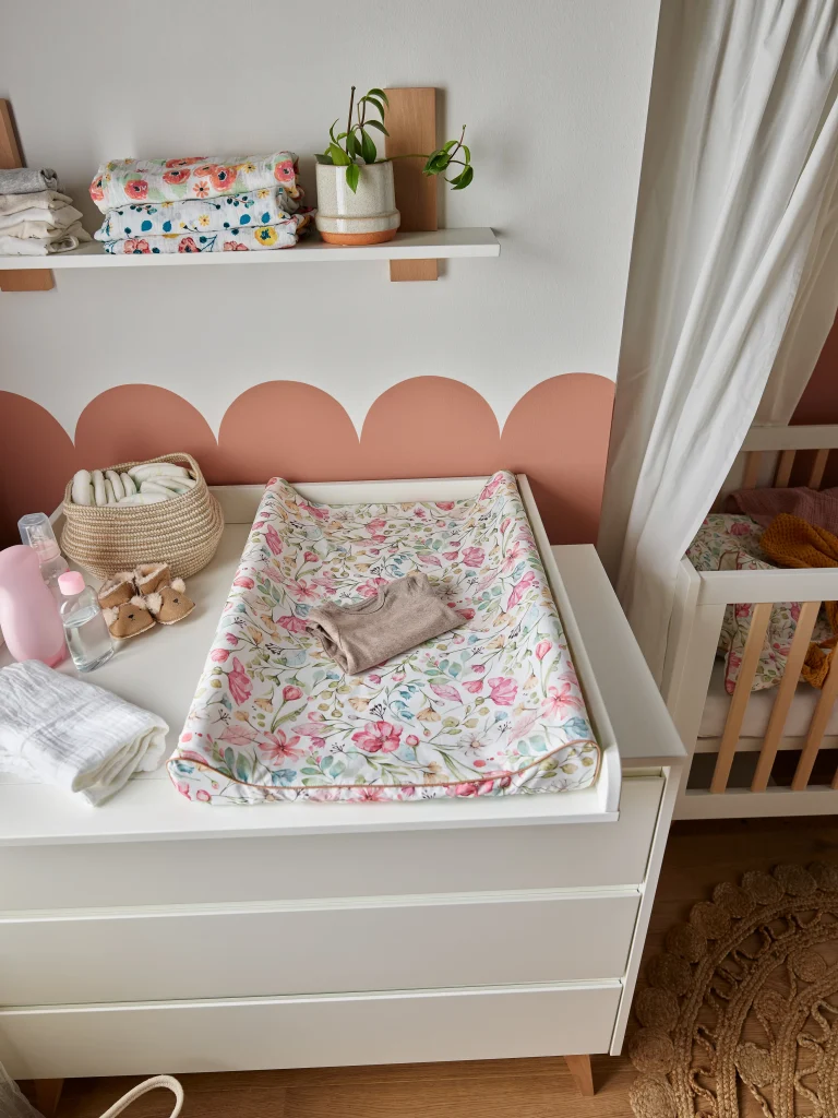 Lifestyle'owe zdjęcie białej komody z szufladami marki Pinio z kolekcji Floral, na blacie znajduje się przewijak pokryty tekstylną matą w kwiatowy wzór, mebel w kolorze białym, standardowy rozmiar, aranżacja w pokoju niemowlęcym
