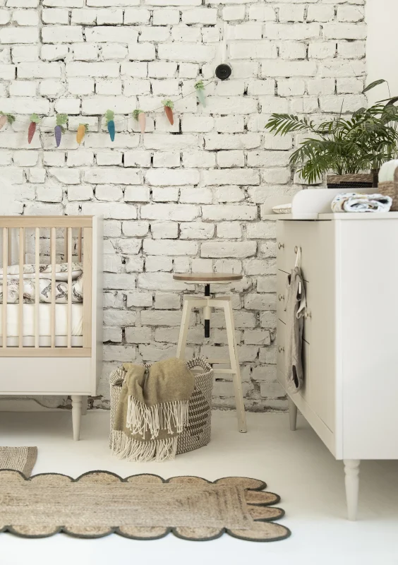 Zdjęcie lifestyle przedstawiające łóżeczko niemowlęce z kolekcji Petite w kolorze szampańskim (jasne drewno), wraz z białą komodą i stołkiem, urządzone w pokoju niemowlęcym z białą ceglaną ścianą, koszem i dywanem