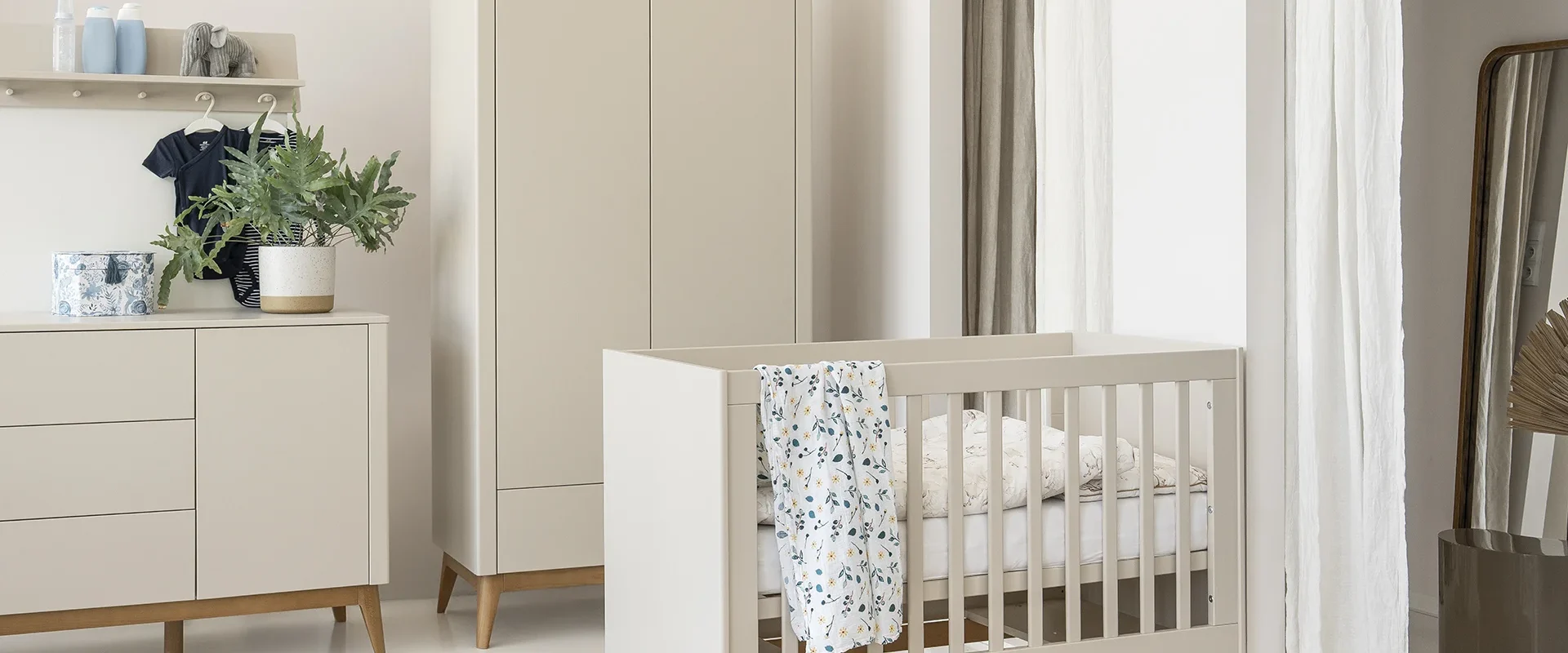 Zdjęcie lifestyle z kolekcji Seth w kolorze champagne przedstawiające łóżeczko niemowlęce z jasnobeżowym korpusem i drewnianymi nóżkami, otoczone meblami z tej samej kolekcji (szafa i komoda)